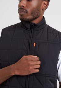 Black quilted vest with high collar and front zipper. Features an orange accent on the chest pocket and smooth, lightweight fabric.