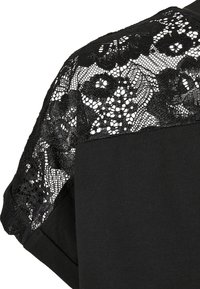 Haut noir à manches courtes, avec une superposition en dentelle noire détaillée sur les épaules. Le tissu a une texture lisse.