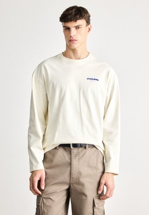 Long-sleeve cream-colored t-shirt with ribbed texture, featuring a blue embroidered logo on the chest. Paired with light brown cargo pants.