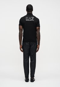 Μαύρο πόλο μπλουζάκι με κοντά μανίκια και λογότυπο EA7 Emporio Armani στην πλάτη, συνδυασμένο με μαύρο υφασμάτινο παντελόνι και μαύρα παπούτσια.