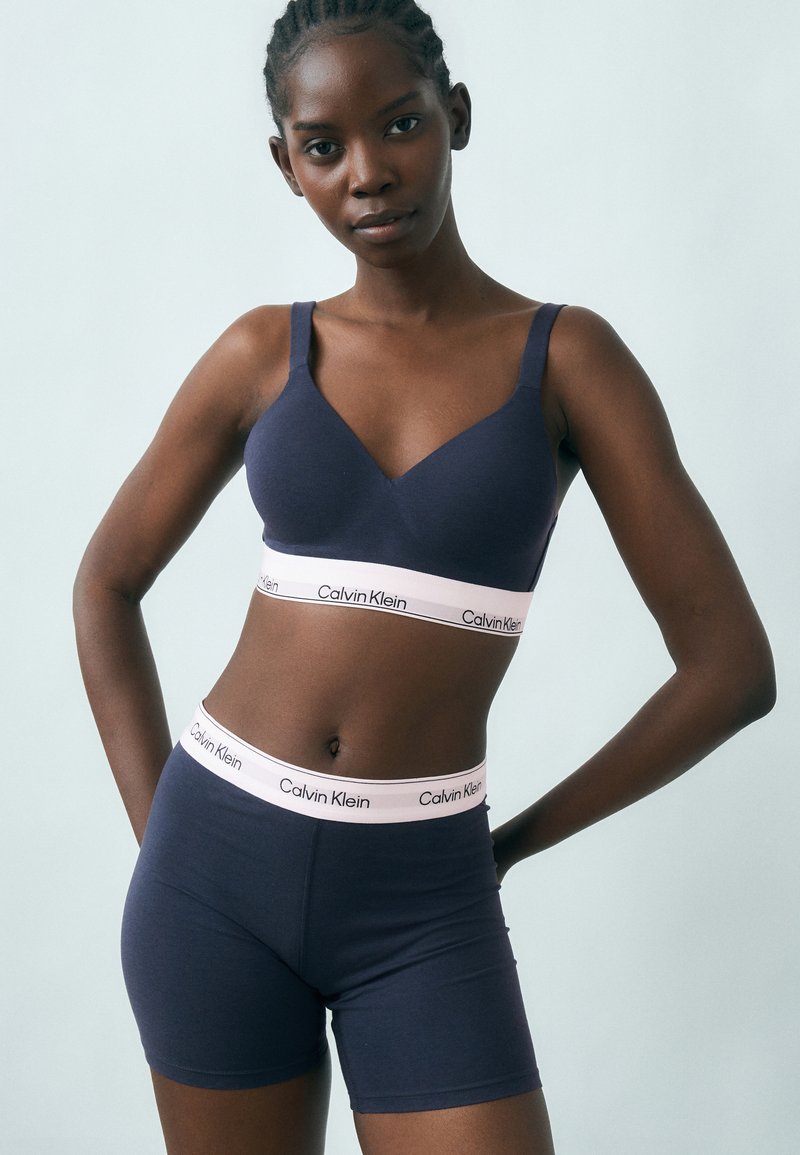 Žena v tmavomodrom bralette značky Calvin Klein a zodpovedajúcich krátkych nohaviciach, stojaca s rukami v bok na svetlomodrom pozadí.