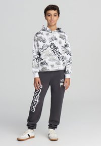 Felpa con cappuccio bianca con motivi graffiti neri e grigi, abbinata a pantaloni della tuta grigi con una grafica bianca e sneakers bianche.