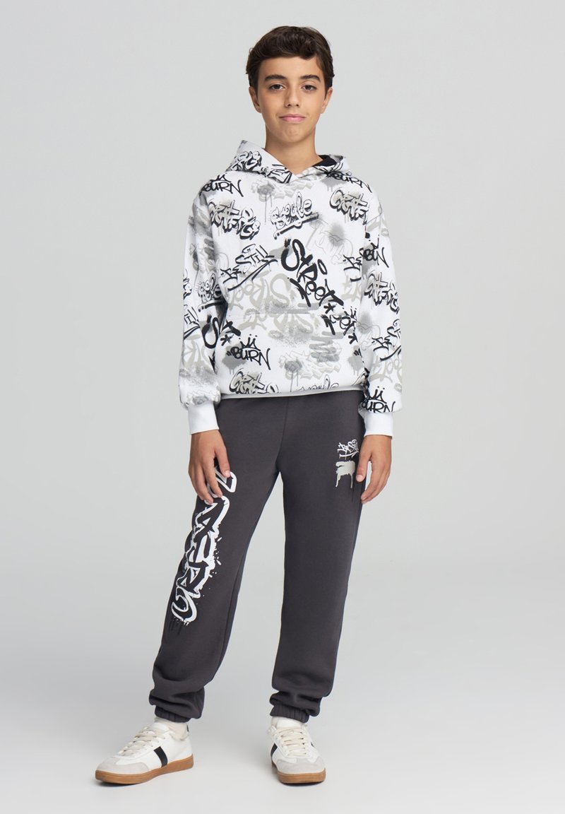 Felpa con cappuccio bianca con motivi graffiti neri e grigi, abbinata a pantaloni della tuta grigi con una grafica bianca e sneakers bianche.