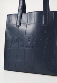 Borsa a tote blu navy realizzata in pelle sintetica testurizzata effetto coccodrillo. Presenta un logo impresso e manici coordinati. Design pulito con forma strutturata.