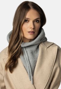 Lys grå strikket hoodie med høj krave under en beige uldfrakke. Bløde teksturer er synlige, og hoodien har snore.