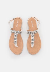 Flat sandals with a beige sole, white strap, and a floral arrangement of clear gemstones on top. Buckle closure on the ankle strap.