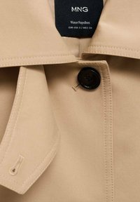 Jachetă bej rezistentă la apă, cu un buton negru mare și detalii cusute, având o guler și o etichetă care afișează informațiile despre marcă și mărime.