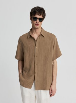 Uomo che indossa una camicia marrone a maniche corte con texture e bottoni, pantaloni bianchi e occhiali da sole neri, in piedi davanti a uno sfondo grigio uniforme.