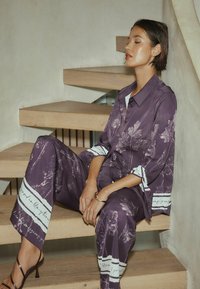 Kvinde med kort mørkt hår sidder på trætrappe iført lilla blomstret skjorte og matchende viddebukser med hælede sandaler.