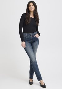 Crna bluza s dugim rukavima i čipkastim obrubom, uparena s tamnoplavim uskim trapericama. Traperice imaju diskretan izblijedjeli uzorak i stilizirane su s crnim šiljastim cipelama.