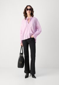 Helle rosa Bluse mit Bindekragen und dekorativer Stickerei, kombiniert mit schwarzen, ausgestellten Jeans und spitzen schwarzen High Heels. Schwarze Handtasche mit Akzenten.