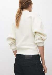 Femme avec queue de cheval châtain clair portant un pull blanc à manches bouffantes et un jean noir taille haute, vue de dos sur un fond uni.