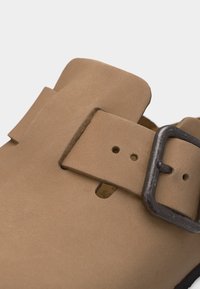 Κοντινό πλάνο ταμπά σουέντ σανδάλι με φαρδιά λωρίδα και σκουρόχρωμη ορθογώνια μεταλλική αγκράφα σε απλό φόντο.