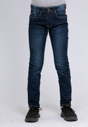 Personne portant un jean skinny bleu foncé et des baskets noires, debout devant un fond clair uni.