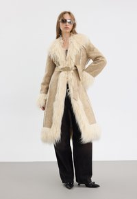 Manteau en fausse fourrure beige avec des manches longues, une ceinture et des bordures duveteuses, associé à un pantalon noir à jambes larges et des escarpins noirs à bout pointu.
