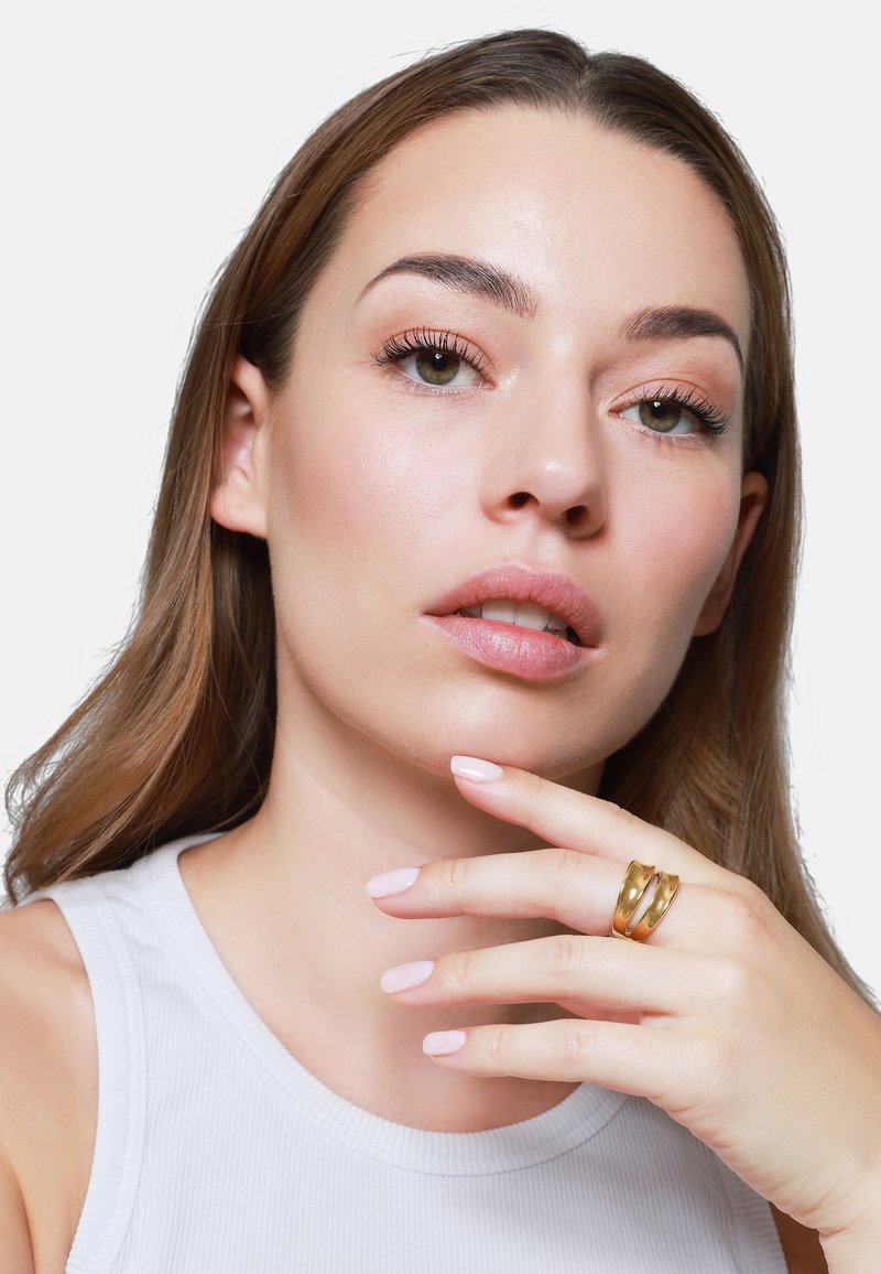 Anillo de oro de tres bandas llevado en una mano con uñas de color rosa claro. El fondo es blanco liso y el cabello del modelo es castaño y ondulado.