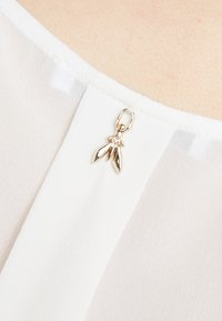 Biała, przezroczysta tkanina z teksturowanym dekoltem, z dodatkiem złotej akcesorii w kształcie liści, przymocowanej do odzieży.