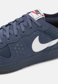 Navy blue sneaker with mesh and suede materials. Features a white Nike swoosh, textured accents, and gray laces. Flat rubber sole.
