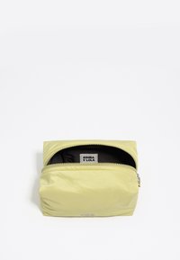 Żółta torba nylonowa z suwakiem, z czarnym wnętrzem i widoczną metką marki. Projekt jest prostokątny z gładką teksturą.