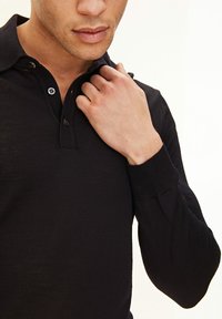 Svart långärmad polo som är tillverkad av ett texturerat stickat tyg, med en två-knapps placket och ribbade ärmslut. Slät yta överlag.