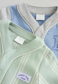Deux ensembles pour bébé : l'un en gris clair et bleu, l'autre en vert avec des rayures blanches. Les deux présentent des fermetures à pression et des étiquettes.