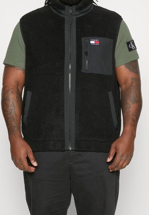 Mann trägt eine schwarze Fleece-Weste mit Reißverschluss über einem grünen Kurzarmshirt mit Markenlogos auf der Brusttasche und dem Ärmel.