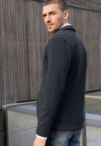 Jonge man in een donkere jas en spijkerbroek die over zijn schouder kijkt, met een houten muur en een metalen reling op de achtergrond.