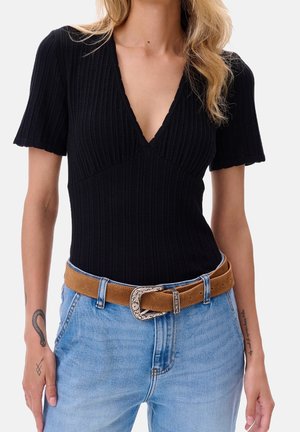 Nainen pukeutuneena mustaan ribbineulottuun V-kaula-aukolliseen lyhythihaiseen toppiin, vaalean sinisiin farkkuihin ja ruskeaan mokkanahkaiseen vyöhön, jonka hopeinen solki on kaiverrettu.