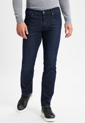 Dunkelblaue Jeans im Slim-Fit, mit fünf Taschen und einem Reißverschluss. Kombiniert mit schwarzen Sneakers mit weißen Sohlen.