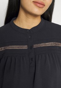 Haut noir avec un col rond et une fermeture à boutons. Il présente des détails décoratifs en dentelle et une texture plissée autour de la taille.