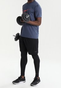 Man som bär sportkläder utför bicepscurl med två svarta hantlar mot en enkel vit bakgrund.