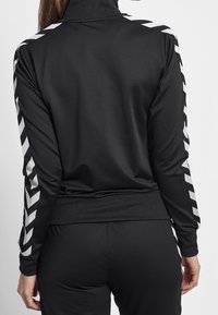 Woman wearing black long-sleeve track jacket and pants with white chevron stripes down sleeves and sides, viewed from the back.