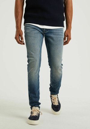 Mężczyzna ubrany w wyblakłe niebieskie dżinsy, granatowy sweter, białą koszulę pod spodem oraz granatowe trampki z kremowymi sznurowadłami, idący na białym tle.