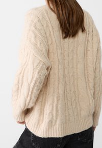 Beige neulepaita, jossa on rakenteellisia kaapelikuviota, ylisuurikokoinen leikkaus, resoroitu helma ja lasketut olkapäät. Pehmeää, pörröistä materiaalia.