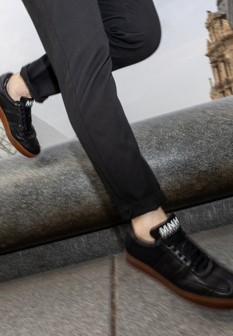 Svarta lädersneakers med en texturerad yta, gummisulor och kontrasterande vit märkning på tungan. Bärs med mörka byxor.