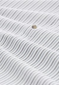 Tela blanca con finas líneas negras onduladas y una sutil textura. Un botón marrón es visible en la superficie, añadiendo un detalle contrastante.