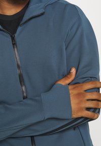 Felpa con zip blu realizzata in un tessuto strutturato, con zip nera e fori per pollice sui polsini aderenti, progettata con un design snello per il massimo comfort.