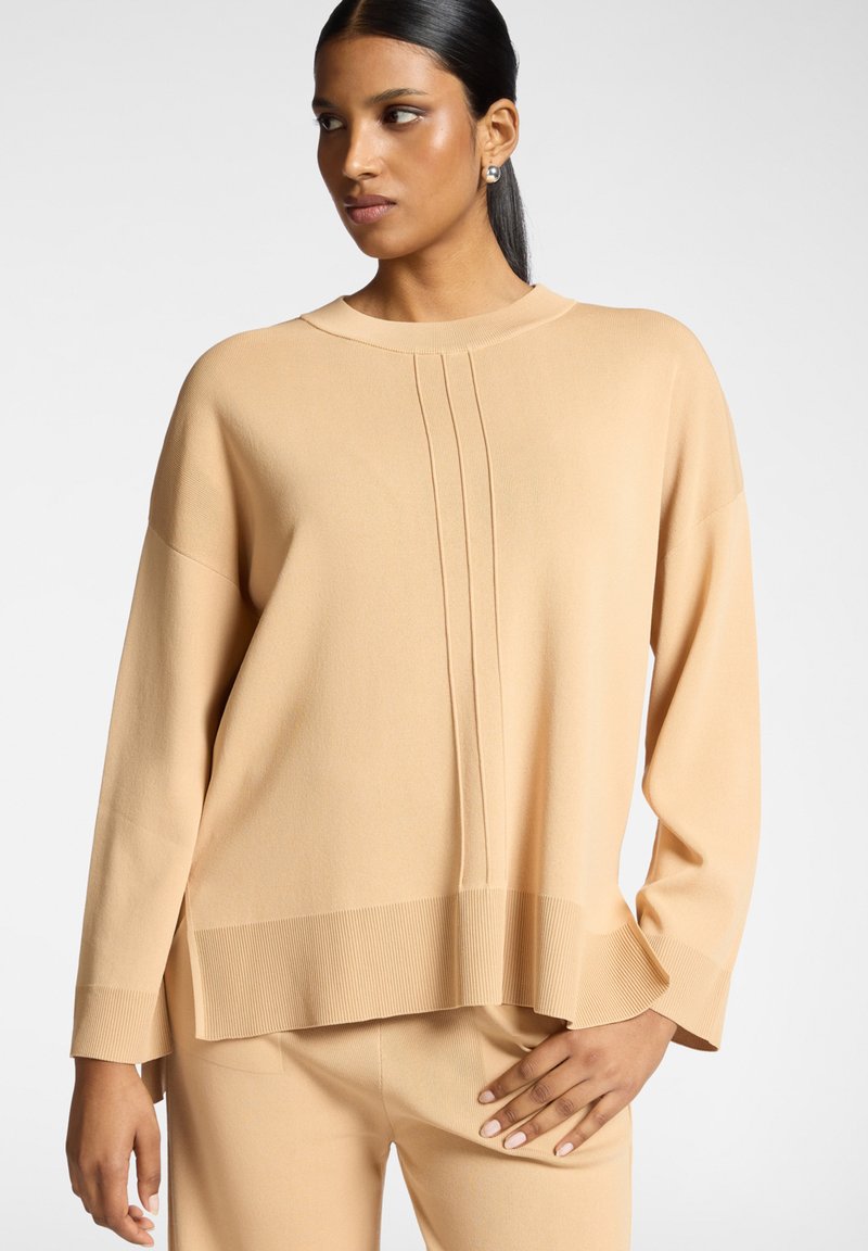 Maglione beige a maniche lunghe con scollo rotondo, caratterizzato da variazioni di trama e pieghe verticali sulla parte anteriore, orlo a coste e vestibilità leggermente oversize.