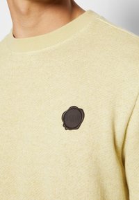 Jasnożółty sweter z teksturowanej tkaniny z ribbowanym dekoltem. Posiada czarną gumową łatę z logo na lewej piersi.