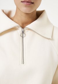 Sudadera cremosa con cremallera y un gran cuello, con una cremallera plateada en la parte delantera y un tejido suave y texturizado.