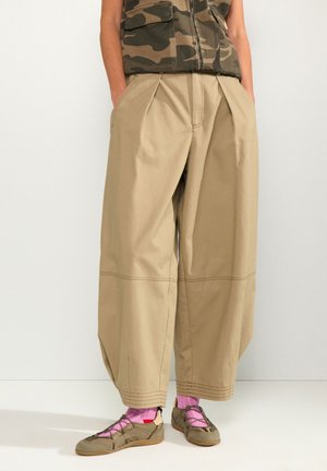 Personne portant un pantalon beige à jambes larges, des chaussettes roses, des chaussures marron décontractées, et une veste sans manches à motif camouflage, les mains dans les poches.