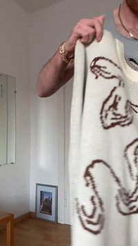 Personne tenant un pull blanc avec des motifs abstraits foncés dans une pièce peu meublée avec une photo encadrée appuyée contre le mur.