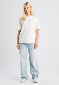 Crèmekleurig katoenen T-shirt met korte mouwen, voorzien van een blauwe grafische print op de borst. In combinatie met lichtblauwe wijde jeans en witte sneakers.