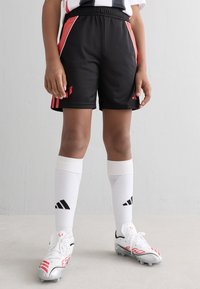 Barn med svarte og røde fotballshorts, hvite Adidas sokker og hvite fotballsko som står på et lyst grått gulv mot en hvit vegg.