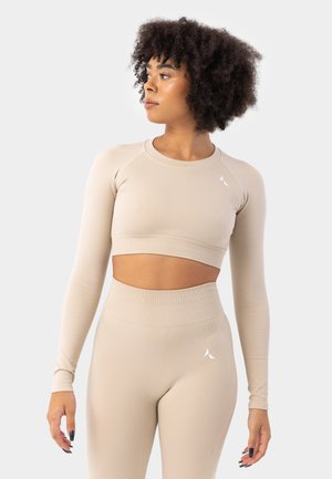 Bežový crop top s dlhým rukávom a ribbed textúrou, kombinovaný s legínami s vysokým pásom. Obe majú na hrudi a na bokoch malé logo.