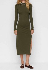 Femme portant une robe midi à manches longues olive avec une fente latérale et des chaussures plates marron, debout devant un fond uni.