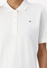 Gros plan d'une personne portant un polo blanc à manches courtes avec un petit logo rouge et bleu sur la poitrine gauche et quatre boutons.