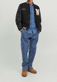 Chaqueta varsity negra con un destacado logo de "N", combinada con una camisa de mezclilla azul clara y jeans de mezclilla holgados. Footwear marrón completa el conjunto.