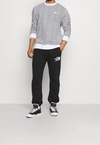 Vit långärmad skjorta med svart prickmönster, tillsammans med svarta sweats som har en vit logotyp. Svarta sneakers med vita snören.