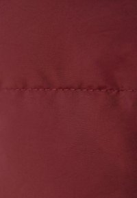 Bordowy materiał o gładkiej teksturze, z eleganckimi detalami szwów. Materiał wydaje się lekki i trwały.